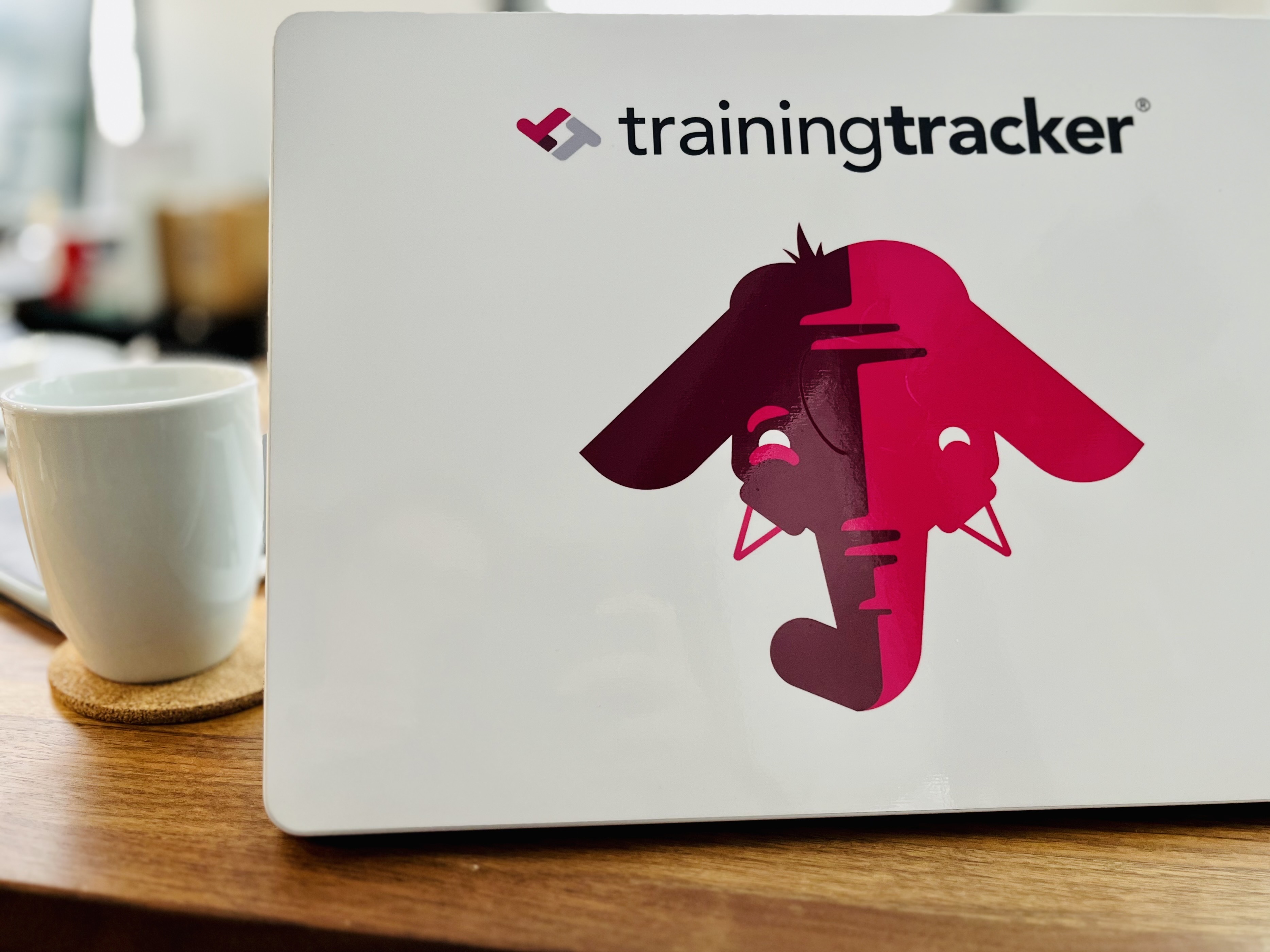 White coffee cup on a cork coaster next to a laptop computer with a Training Tracker branded cover.