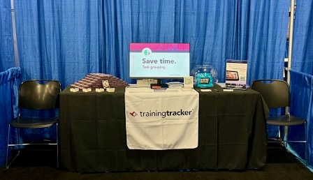 A trade show booth for 'trainingtracker' with a black tablecloth and logo banner draped in front. The table displays promotional materials, including what appears to be pamphlets or brochures, a monitor with 'Save time' messaging, and some branded merchandise. Two chairs are positioned in front of the table, with a blue curtain backdrop. The setup is compact and professional, designed for conference networking.