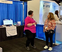 At a trade show or conference, two women are engaged in conversation in front of exhibition booths. One woman in a bright pink top stands talking with another woman in a patterned blouse. Behind them are vendor displays with blue draping and signage, including what appears to be the 'trainingtracker' booth visible on the left with its tablecloth and display materials.