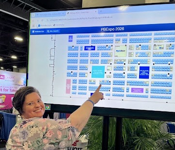 A person in a floral patterned shirt points at a large digital display showing a convention floor map labeled 'PBEpro 2026'. The screen displays a color-coded grid layout of exhibition booths and spaces. The individual appears to indicate a specific location on the map, likely helping with navigation at the trade show or conference.