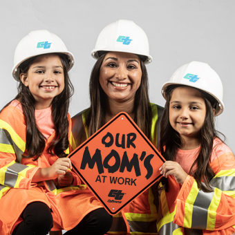 Image shows three people in construction safety gear - an adult woman in the center with two young girls beside her. All wear hard hats and high-visibility orange vests with reflective stripes. They're smiling while holding an orange diamond-shaped sign reading "OUR MOM'S AT WORK" with a construction company logo. The photo appears to celebrate women in construction/infrastructure roles.
