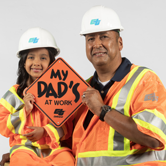 Image shows a man and a young girl in construction safety gear, including white hard hats and high-visibility orange vests with reflective stripes. They're smiling at the camera while holding an orange diamond-shaped sign that reads "MY DAD'S AT WORK" with a construction company logo. The photo appears to celebrate family connections to construction/infrastructure work and workplace safety.
