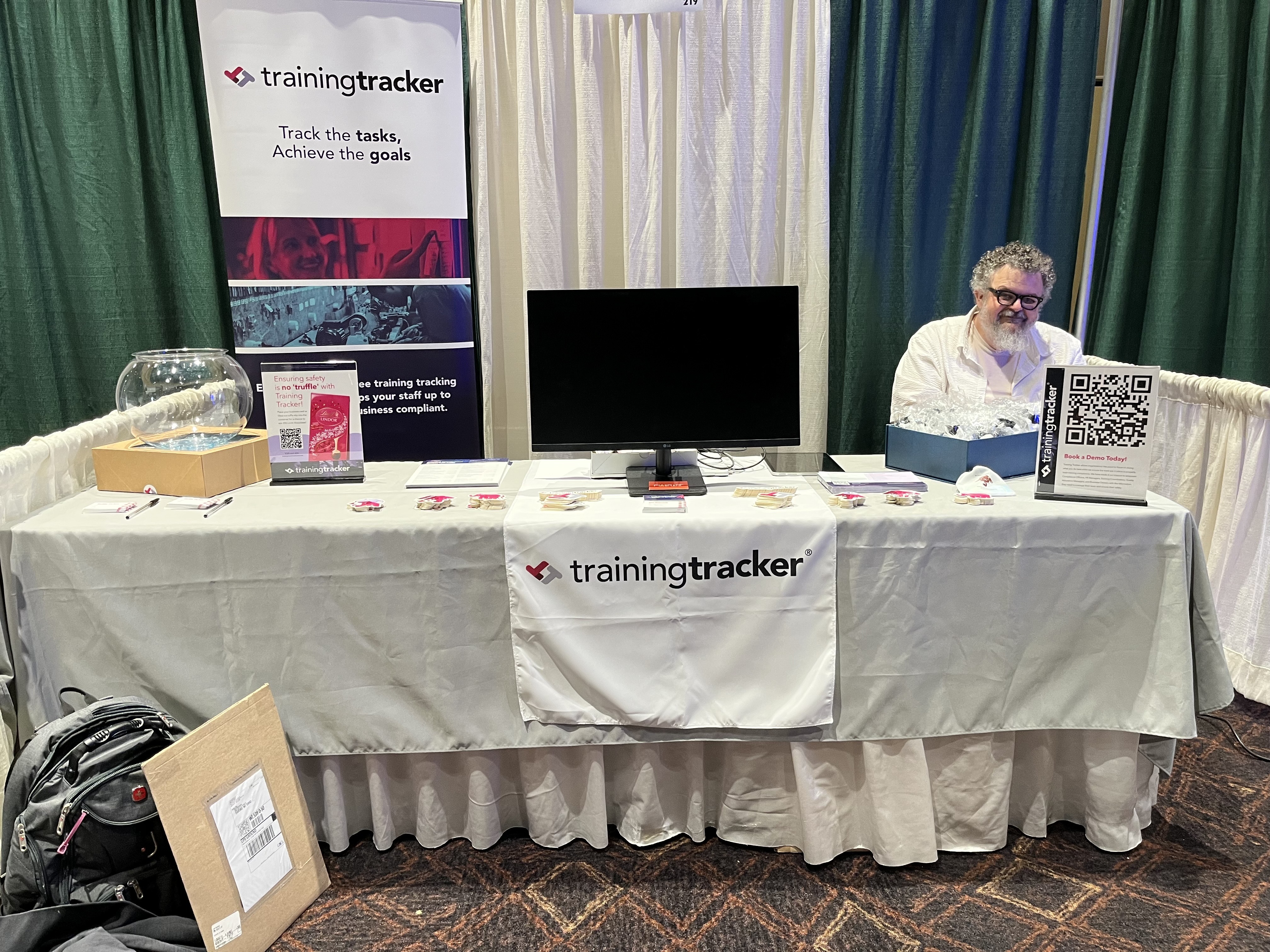 Exhibition booth with TrainingTrainer logo display. Man sitting at table with promotional materials and literature.
