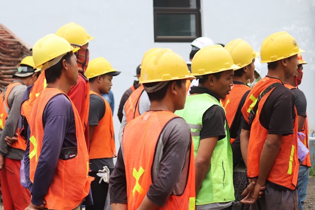 Construction workers wearing yellow hard hats and bright orange and green high-visibility safety vests gathered together at a job site, with buildings visible in the background.