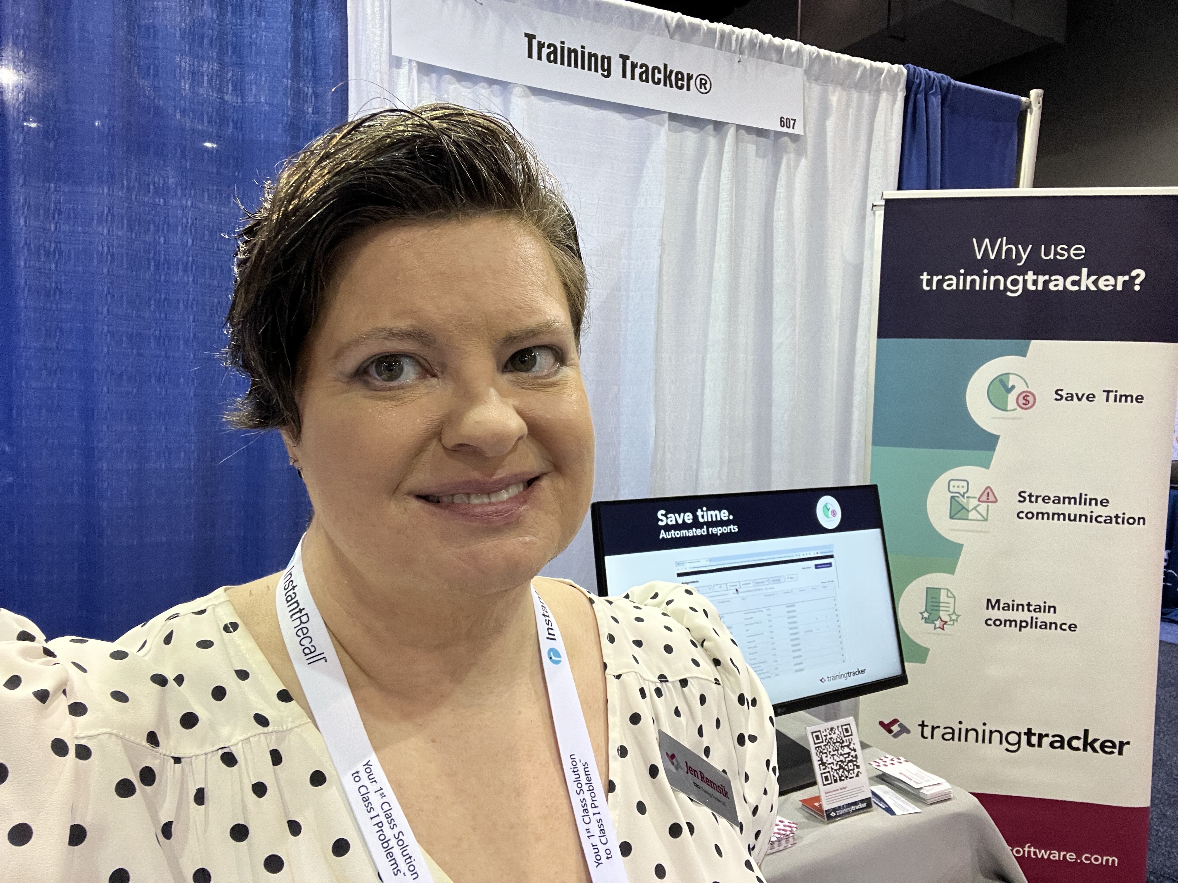 A woman in a polka dot blouse smiles at a trade show booth for Training Tracker®. The booth displays the software's benefits including "Save Time," "Streamline communication," and "Maintain compliance," with a tablet showing the interface and promotional materials on the counter.