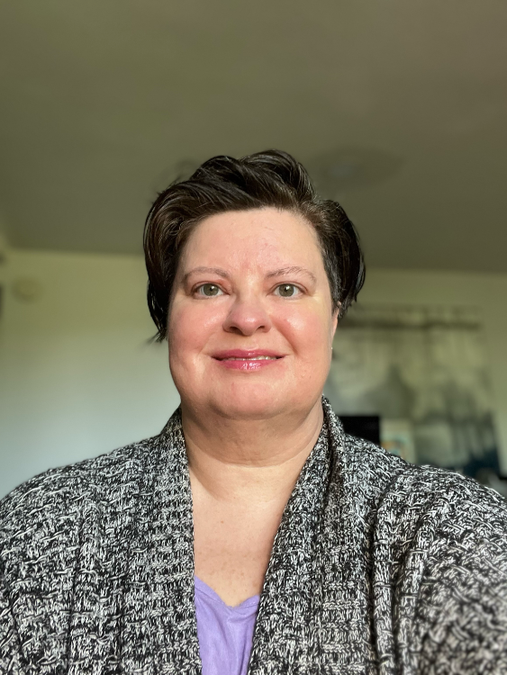 A person with short dark hair smiling at the camera, wearing a gray and white marled cardigan over a purple top, photographed indoors with a neutral background.
