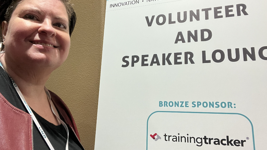 Professional woman with short dark hair wearing a lanyard and burgundy jacket smiles at the camera while standing next to a conference sign reading "VOLUNTEER AND SPEAKER LOUNGE" with "BRONZE SPONSOR: trainingtracker" displayed below. The setting appears to be at an innovation or professional development event, with a beige wall visible in the background.